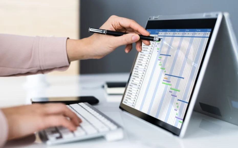Person uses a pen to point at a calendar-style schedule displayed on a tilted touchscreen monitor while typing on a keyboard nearby.