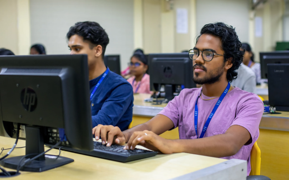 Young man with glasses typing at a desktop computer in a classroom computer lab, others working at screens in the background.