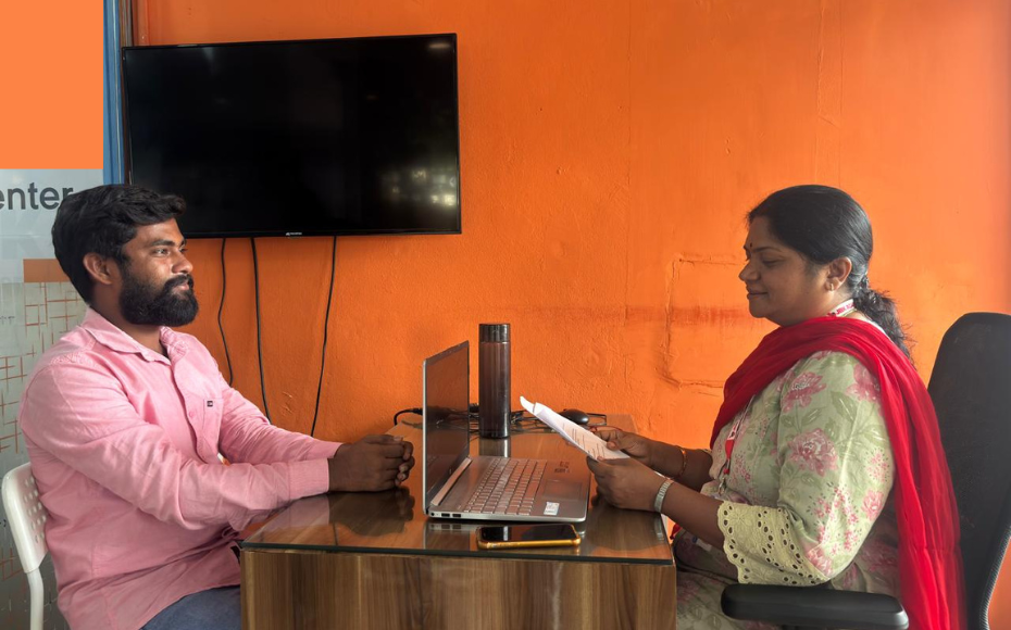 Man in a pink shirt sits across a desk from a woman in a red scarf, holding papers as she types on a laptop in a bright orange office.