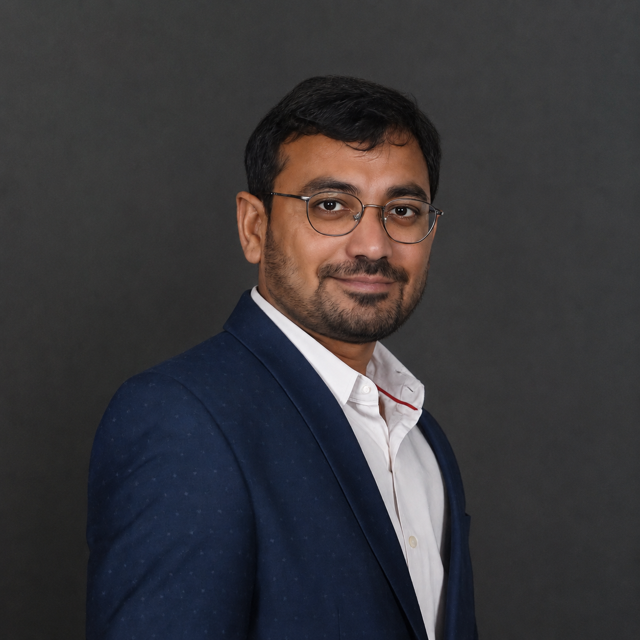 Professional headshot of a man in a navy suit and white shirt with glasses, slight smile, neutral dark background