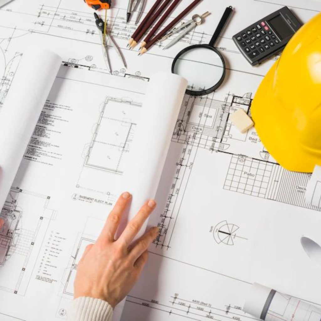 Architectural blueprints on a desk with drafting tools, pencils, magnifying glass, calculator, and a yellow hard hat nearby.