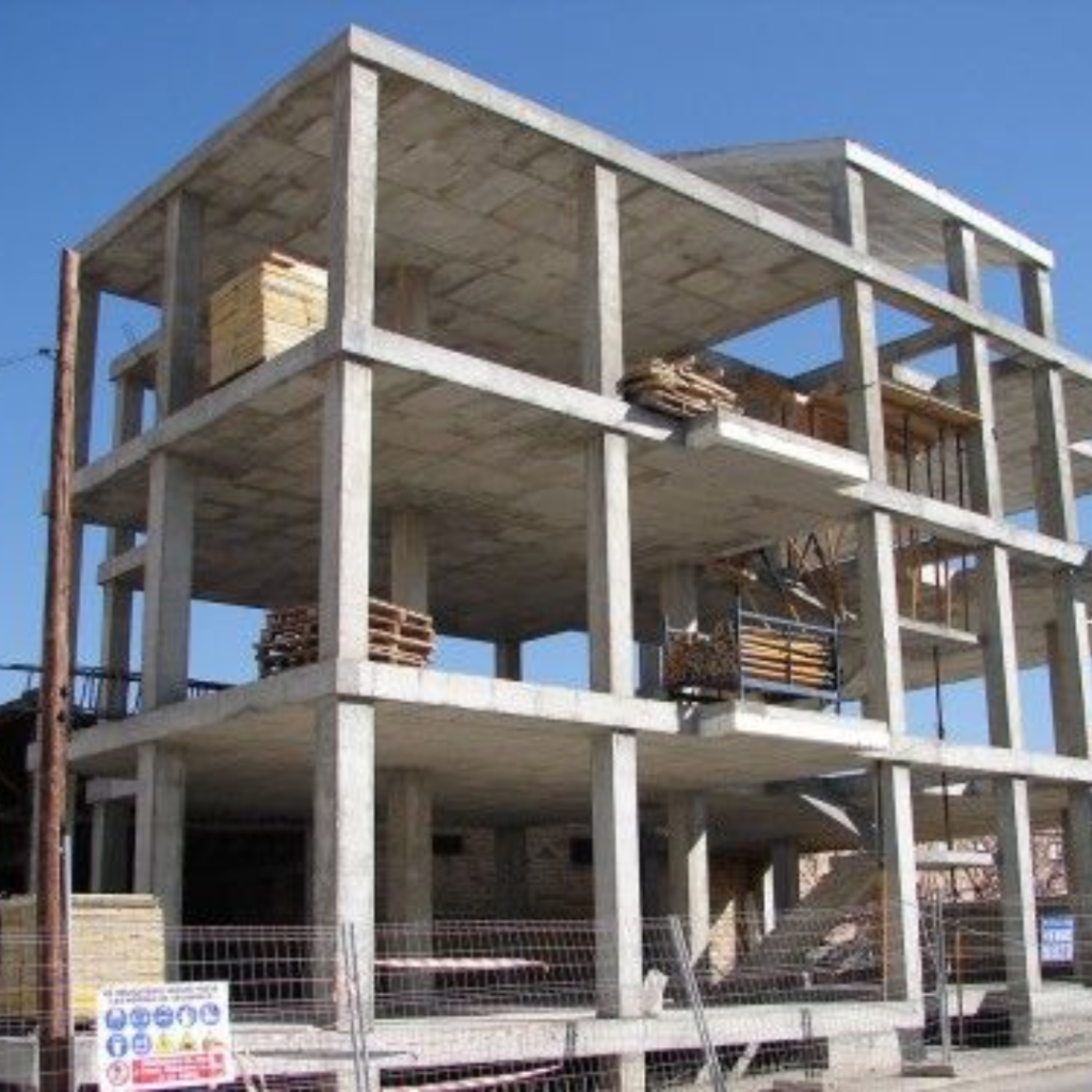 Concrete-framed building under construction with exposed floors and stacked pallets on the upper levels under a clear blue sky