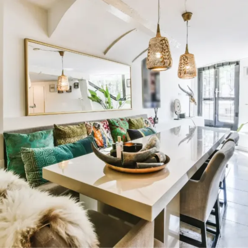 Bright, modern dining area with a long white table, beige chairs, and colorful patterned cushions along a bench under a large wall mirror.
