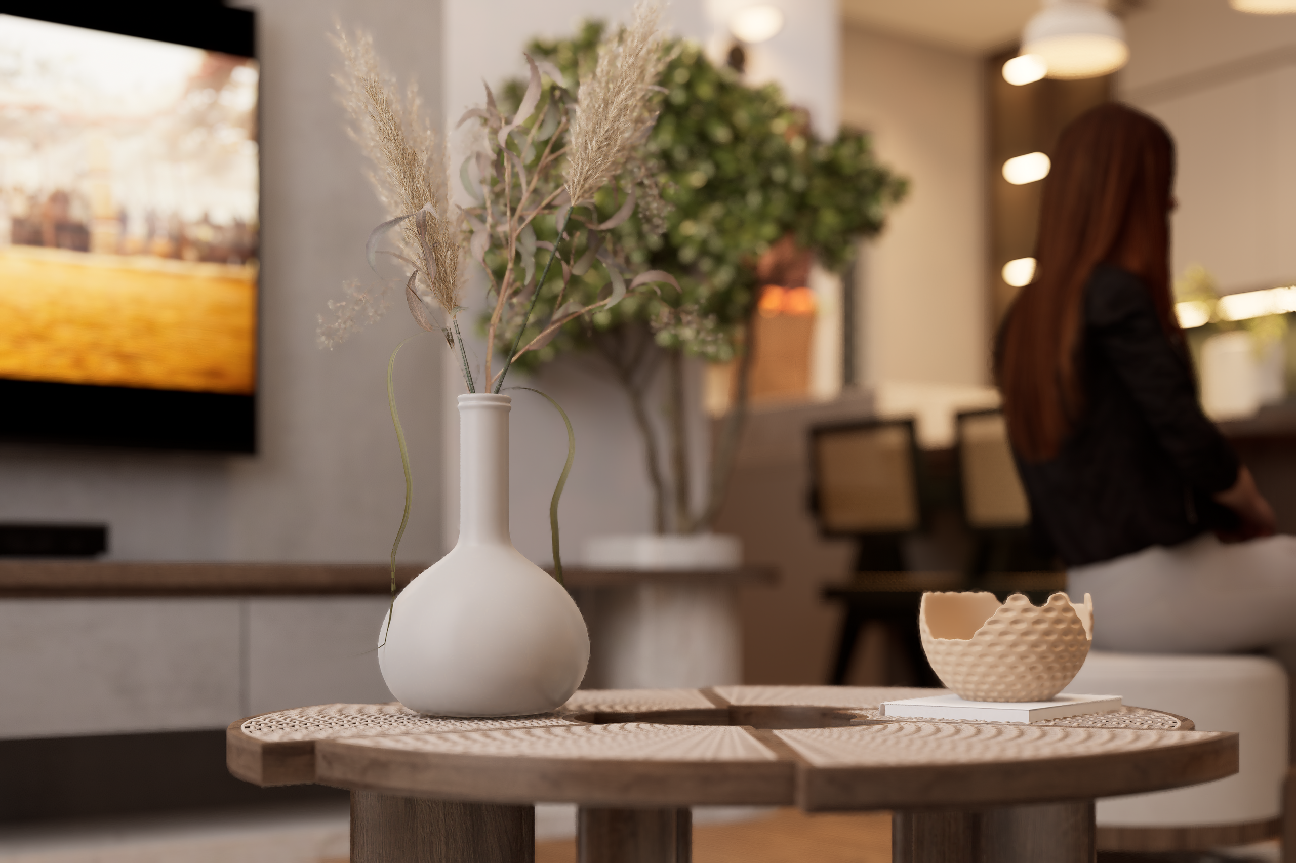 White ceramic vase with dried grasses on a round wooden table in a modern lounge area. It’s softly lit with a blurred background and a person standing/working in the distance.